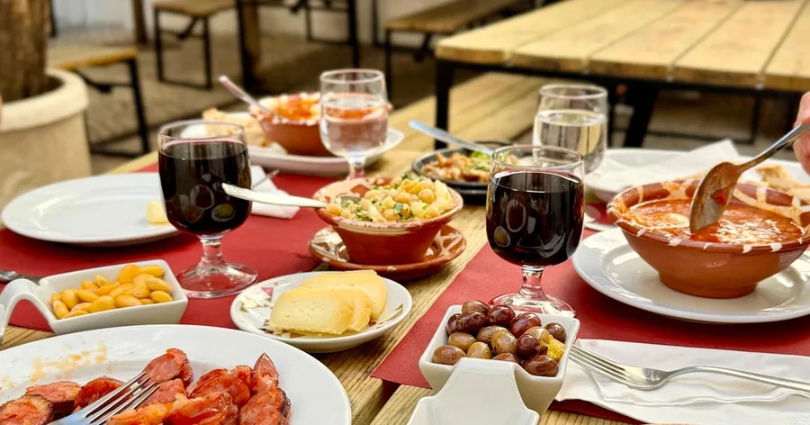 Uma mesa de tasca com vinho tinto, chouriço assado, arroz, queijo e pequenos pratos